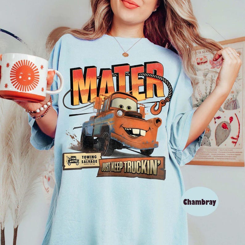 Puede incluir: Camiseta azul claro con un dise&ntilde;o de Mate de la pel&iacute;cula Cars, con la palabra "MATER" en naranja y amarillo. La camiseta tambi&eacute;n tiene el texto "JUST KEEP TRUCKIN'" y una taza con un sol sonriente.
