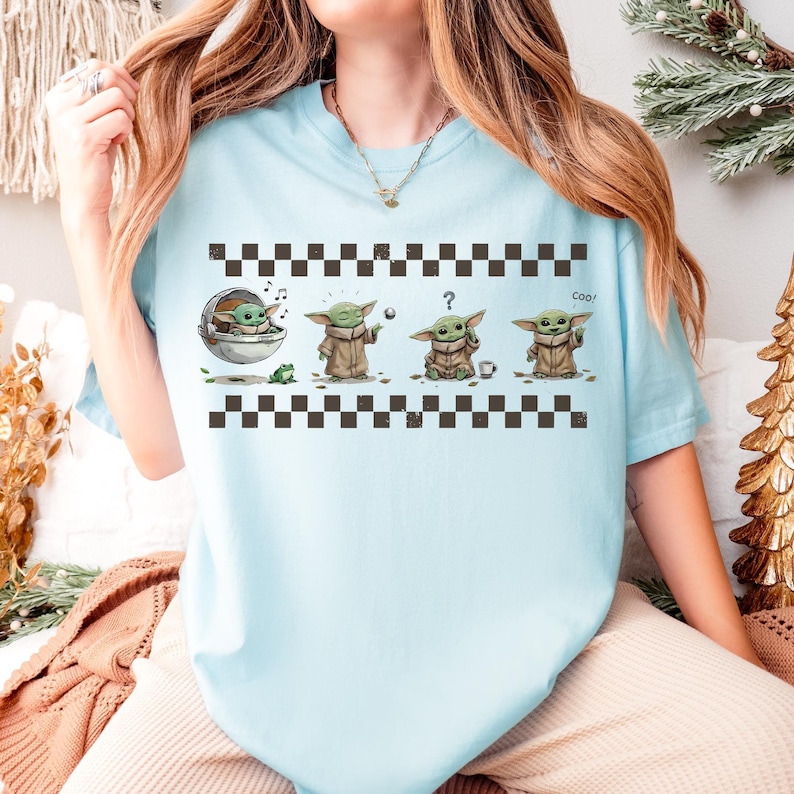 May include: Light blue t-shirt featuring a graphic of a character in various poses. The character is green with large ears and is wearing a brown robe. The shirt has a black and white checkered pattern at the top and bottom. The word "Cool" is printed on the shirt.