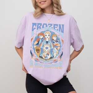 May include: A light purple t-shirt featuring the word "FROZEN" in blue, with a circular graphic of characters from the movie. The graphic includes Elsa, Anna, Kristoff, Olaf, and Sven. The text "IN THIS TOGETHER" is below the graphic.