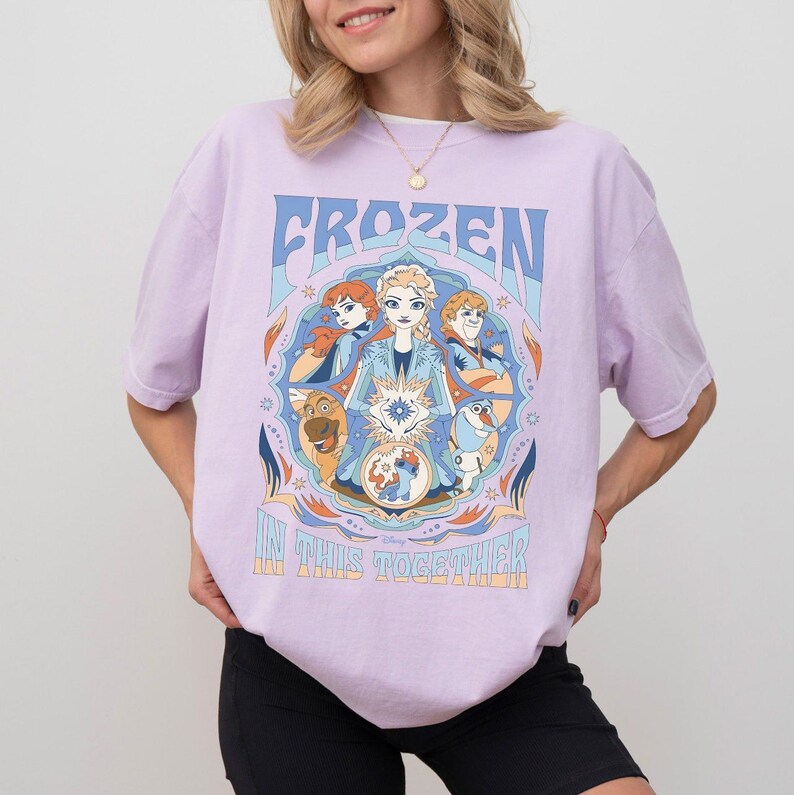 May include: A light purple t-shirt featuring the word "FROZEN" in blue, with a circular graphic of characters from the movie. The graphic includes Elsa, Anna, Kristoff, Olaf, and Sven. The text "IN THIS TOGETHER" is below the graphic.