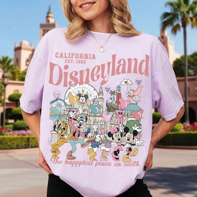 May include: A lilac t-shirt with a Disneyland graphic. The design includes the text "California Est. 1955 Disneyland" and Disney characters. The phrase "The happiest place on earth" is also included on the shirt.