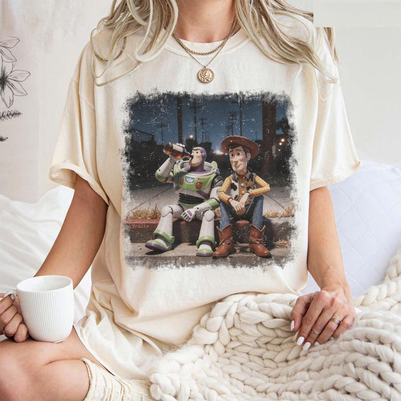 Puede incluir: Camiseta color crema con un gr&aacute;fico desgastado de Buzz Lightyear y Woody de Toy Story. Buzz est&aacute; bebiendo de una botella, mientras que Woody est&aacute; sentado a su lado. El gr&aacute;fico est&aacute; enmarcado con un borde oscuro, creando un aspecto vintage.