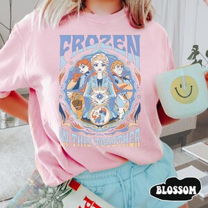 May include: Pink t-shirt featuring a colorful graphic from the Disney movie Frozen. The design includes the text "FROZEN" and "IN THIS TOGETHER" with images of the characters Elsa, Anna, Kristoff, Olaf, and Sven. The shirt is a light pink color.
