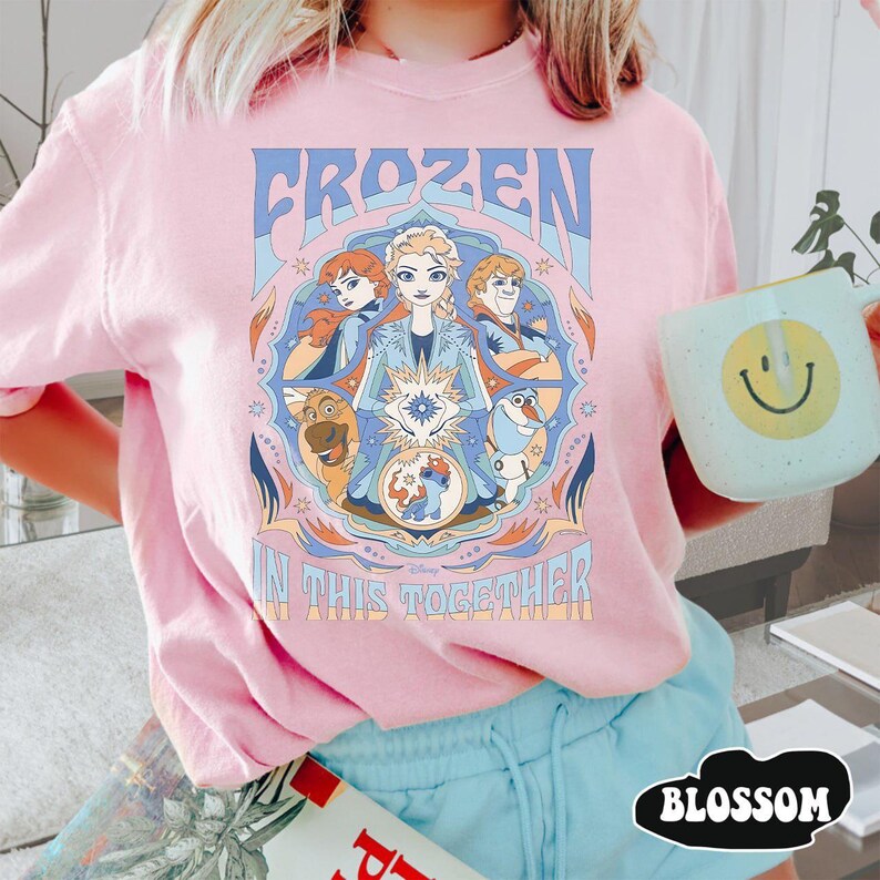 May include: Pink t-shirt featuring a colorful graphic from the Disney movie Frozen. The design includes the text "FROZEN" and "IN THIS TOGETHER" with images of the characters Elsa, Anna, Kristoff, Olaf, and Sven. The shirt is a light pink color.