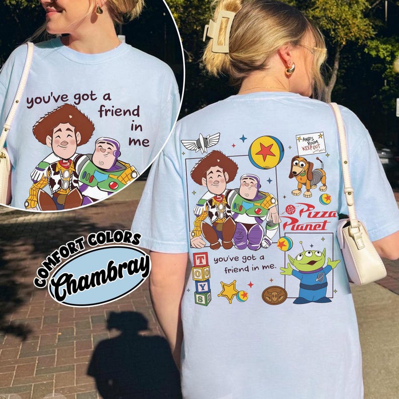 Op de afbeelding: Lichtblauw Comfort Colors Chambray T-shirt met een afbeelding van Woody en Buzz Lightyear en de tekst "you've got a friend in me". De achterkant van het shirt bevat extra Toy Story-personages en het Pizza Planet-logo.
