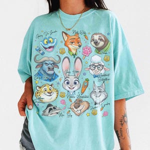 K&ouml;nnte beinhalten: Ein hellblaues T-Shirt mit Zeichentrickfiguren aus Zootopia. Das Design umfasst Judy Hopps, Nick Wilde, Flash und andere Charaktere mit ihren Namen. Das Shirt hat einen Rundhalsausschnitt und kurze &Auml;rmel.