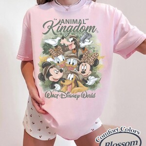 May include: A light pink t-shirt featuring a graphic of Disney characters, including Mickey and Minnie Mouse, Donald and Daisy Duck, Goofy, and Pluto. The shirt has the words "Animal Kingdom" and "Walt Disney World" printed on it. The shirt also has the words "Comfort Colors Blossom" printed on it.