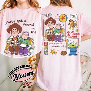 Op de afbeelding: Lichtroze t-shirt met een cartoon illustratie van Woody en Buzz Lightyear en de tekst "you've got a friend in me". De achterkant van het shirt heeft het Pizza Planet logo en andere karakter graphics.