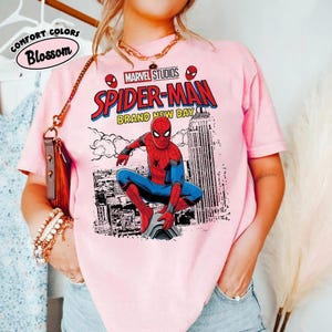 Pode incluir: Camiseta rosa claro com um gr&aacute;fico do Homem-Aranha da Marvel Studios. O design inclui o Homem-Aranha em um traje vermelho e azul, o texto "SPIDER-MAN BRAND NEW DAY" e o logotipo da Marvel Studios. A camisa tamb&eacute;m tem o texto "Comfort Colors Blossom" em um c&iacute;rculo.
