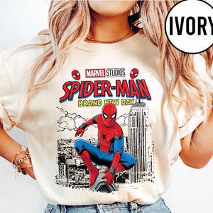 Pode incluir: Camisola de cor marfim com um design de banda desenhada do Homem-Aranha. O gr&aacute;fico inclui o log&oacute;tipo da Marvel Studios, as palavras "Spider-Man Brand New Day" e uma ilustra&ccedil;&atilde;o do Homem-Aranha. A camisola tem gola redonda.