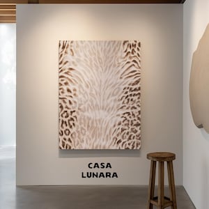 Könnte beinhalten: Ein rechteckiges Wandkunstwerk mit einem braun-weißen Tiermuster mit leopardenartigen Flecken. Unter dem Kunstwerk ist der Text CASA LUNARA in fetten schwarzen Buchstaben gedruckt. Ein hölzerner Hocker mit vier Beinen steht auf der rechten Seite.