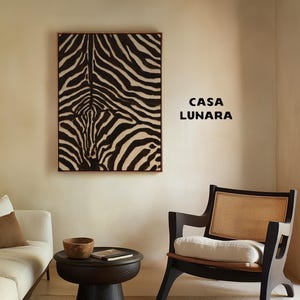 May include: A rectangular wall art piece featuring a black and white zebra stripe pattern in a thin wooden frame, positioned next to the black text CASA LUNARA on a beige wall, above a black pedestal table and a black chair with cane backing.