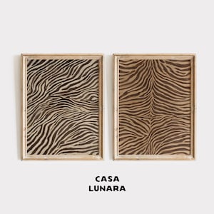 Puede incluir: Dos láminas de arte mural rectangulares en marcos de madera clara, cada una con un patrón de rayas de cebra en marrón oscuro y tostado. El texto CASA LUNARA está impreso en letras mayúsculas negras bajo los dos cuadros sobre fondo blanco.