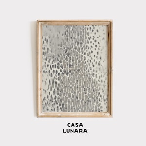 Puede incluir: Una impresión de arte mural rectangular en un marco de madera clara con un patrón abstracto de formas orgánicas y alargadas en gris y blanco que recuerdan a una piel de animal texturizada. El texto CASA LUNARA aparece impreso bajo el marco.