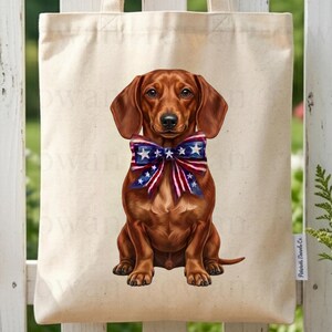 May include: A beige canvas tote bag featuring a brown dachshund wearing a patriotic bow tie with red, white, and blue stars and stripes. The bag is hanging on a white picket fence with an American flag in the background.