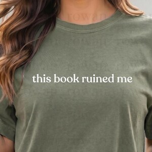 Op de afbeelding: Olijfgroen T-shirt met de witte tekst "this book ruined me". Het shirt heeft een ronde hals en korte mouwen. De stof lijkt zacht en comfortabel, geschikt voor vrijetijdskleding. De tekst staat in het midden op de voorkant van het shirt.