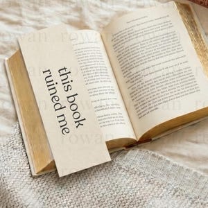 May include: An open book with gold-edged pages and text. A bookmark with the words "this book ruined me" is inserted. The book rests on a textured, light-colored surface, with other books visible in the background.