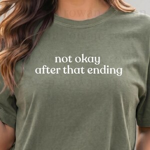 May include: Olive green t-shirt with the text "not okay after that ending" in a white, sans-serif font. The shirt has a crew neck and short sleeves. The fabric appears to be soft and slightly textured.