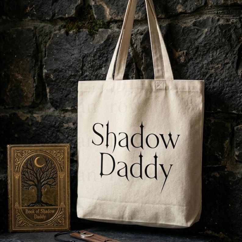 May include: A beige canvas tote bag with the words "Shadow Daddy" printed in black. Beside the bag is a book with a tree design and the text "Book of Shadow Daddy". The background is a dark stone wall.