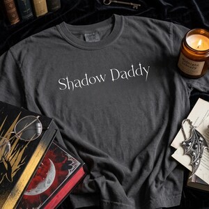 May include: Dark gray t-shirt with the text "Shadow Daddy" in a white, stylized font. The shirt is on a dark surface with books, a candle, and a decorative pendant.