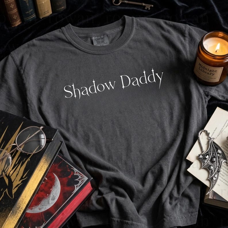 May include: Dark gray t-shirt with the text "Shadow Daddy" in a white, stylized font. The shirt is on a dark surface with books, a candle, and a decorative pendant.