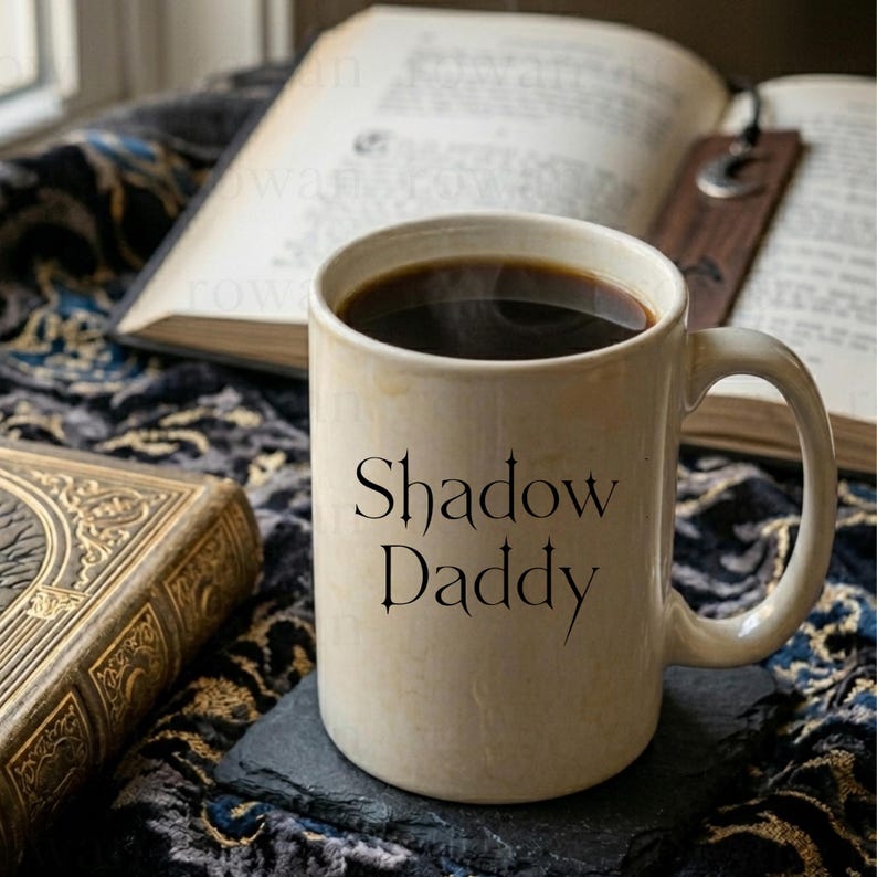 May include: A beige ceramic mug filled with dark coffee, featuring the text "Shadow Daddy" in black gothic lettering. The mug sits on a dark coaster, with an open book and a decorative book in the background.