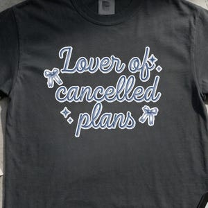 May include: Dark gray t-shirt with the text "Lover of cancelled plans" in a blue script font. The text is outlined in white and features decorative bow and star accents. A casual, comfortable tee.