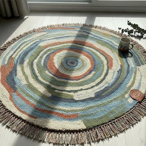 Puede incluir: Una alfombra redonda con un diseño concéntrico en tonos azules, verdes, naranjas y crema. La alfombra tiene un borde con flecos y un patrón de anillos de árbol. Una taza y una etiqueta de cuero están sobre la alfombra.