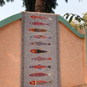 May include: A rectangular gray textile wall hanging featuring a vertical row of ten stylized fish in various colors and patterns. The piece has a dark geometric border and short white fringe at the top and bottom edges, displaying a woven texture.