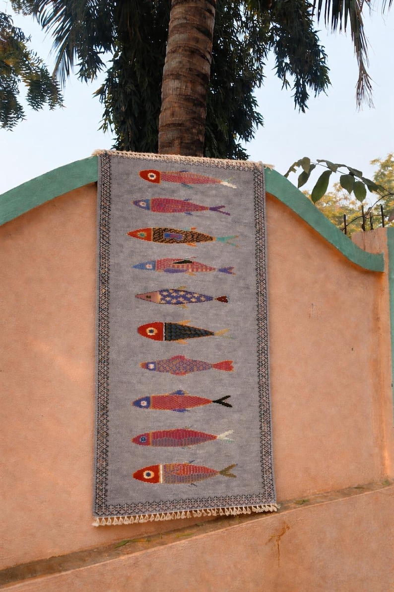 May include: A rectangular gray textile wall hanging featuring a vertical row of ten stylized fish in various colors and patterns. The piece has a dark geometric border and short white fringe at the top and bottom edges, displaying a woven texture.