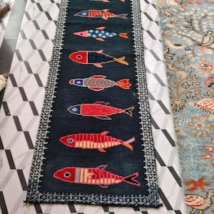 May include: A long, rectangular dark blue runner rug featuring a vertical row of nine stylized fish in various colors including red, orange, blue, and pink. The rug is framed by a white and black geometric border pattern along all four edges.