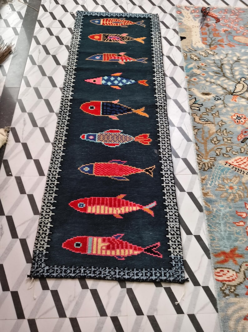 May include: A long, rectangular dark blue runner rug featuring a vertical row of nine stylized fish in various colors including red, orange, blue, and pink. The rug is framed by a white and black geometric border pattern along all four edges.