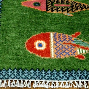 May include: A green area rug featuring two stylized fish motifs in red, orange, and blue. The rug has a dark blue patterned border and white fringe along the bottom edge. The texture appears tufted with a dense pile throughout the surface.