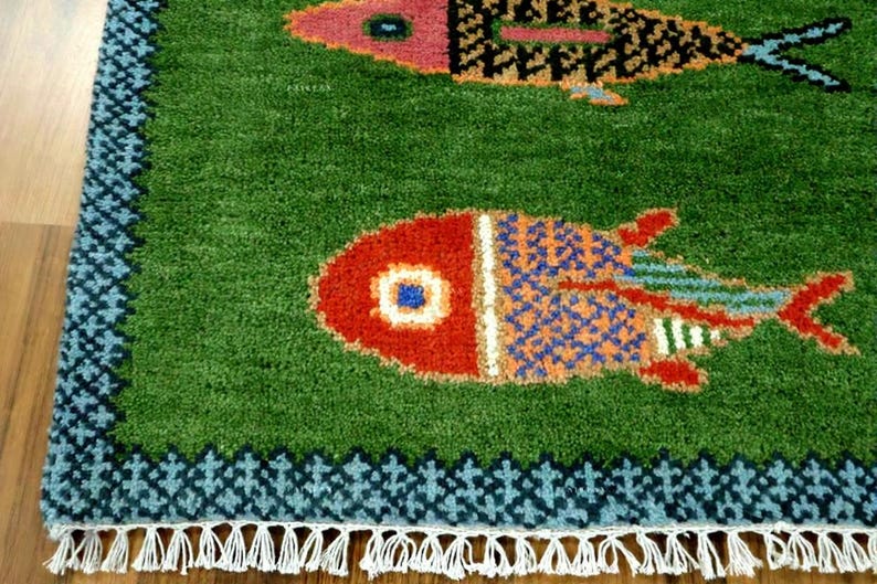 May include: A green area rug featuring two stylized fish motifs in red, orange, and blue. The rug has a dark blue patterned border and white fringe along the bottom edge. The texture appears tufted with a dense pile throughout the surface.