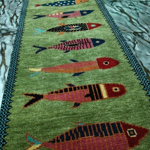 May include: A long, rectangular green runner rug featuring a repeating pattern of stylized fish in various colors including red, pink, blue, and orange. The rug has a dark blue and black patterned border along both long edges of the textile.