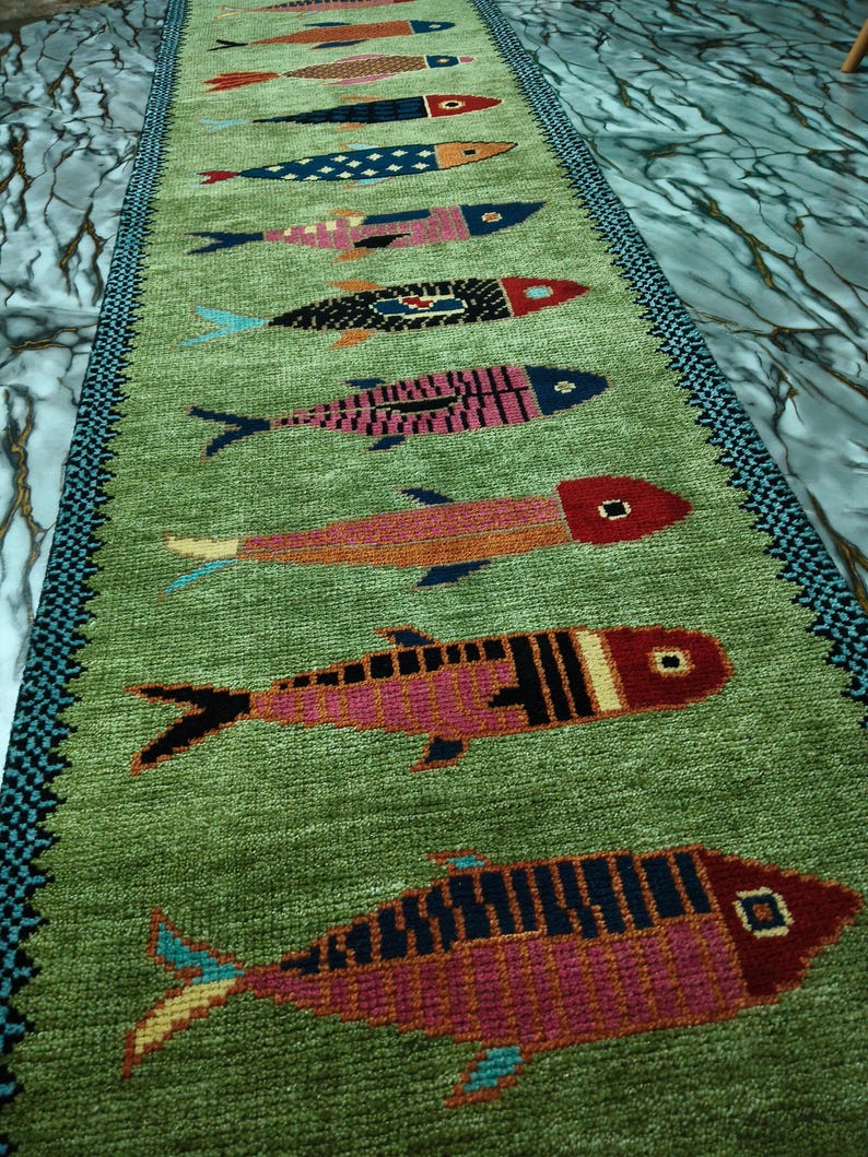 May include: A long, rectangular green runner rug featuring a repeating pattern of stylized fish in various colors including red, pink, blue, and orange. The rug has a dark blue and black patterned border along both long edges of the textile.