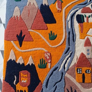 May include: A tufted rug featuring a landscape design with orange fields, blue mountains, a winding blue river, and small houses. The scene includes a yellow lion figure, green shrubbery, and a light blue sky, all rendered in a textured, multi-colored textile.