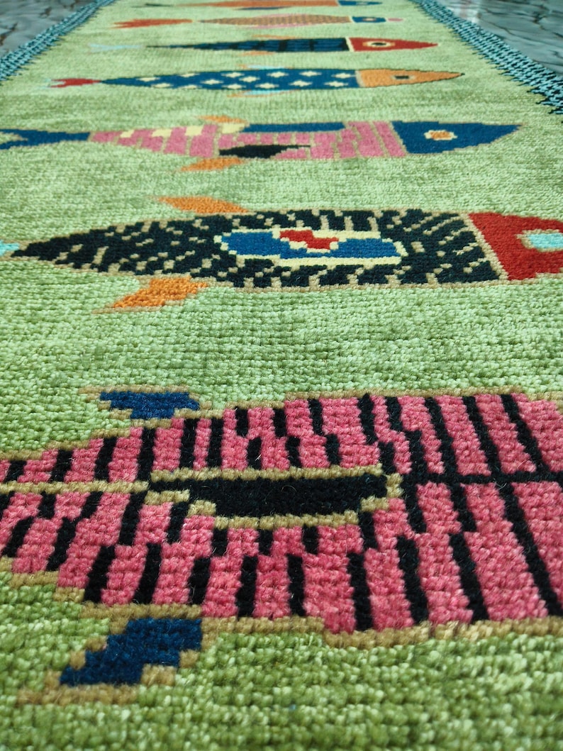 May include: A rectangular runner rug with a light green textured background featuring a repeating pattern of stylized fish in various colors including pink, black, blue, and orange, finished with a dark blue border along the long edges.