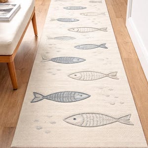 Peut inclure: Tapis de couloir crème avec un motif répété de poissons stylisés dans des tons de gris. Le tapis a une bordure à franges et est posé sur un sol en bois. Le motif de poissons ajoute un thème nautique ou côtier à la décoration.