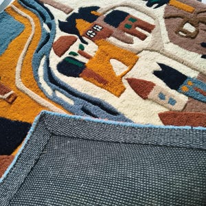 May include: A tufted rug featuring a cream background with a colorful landscape design including a yellow sun, blue river, and brown buildings. The rug has a textured pile surface and a dark gray, grid-patterned non-slip backing visible at the bottom.