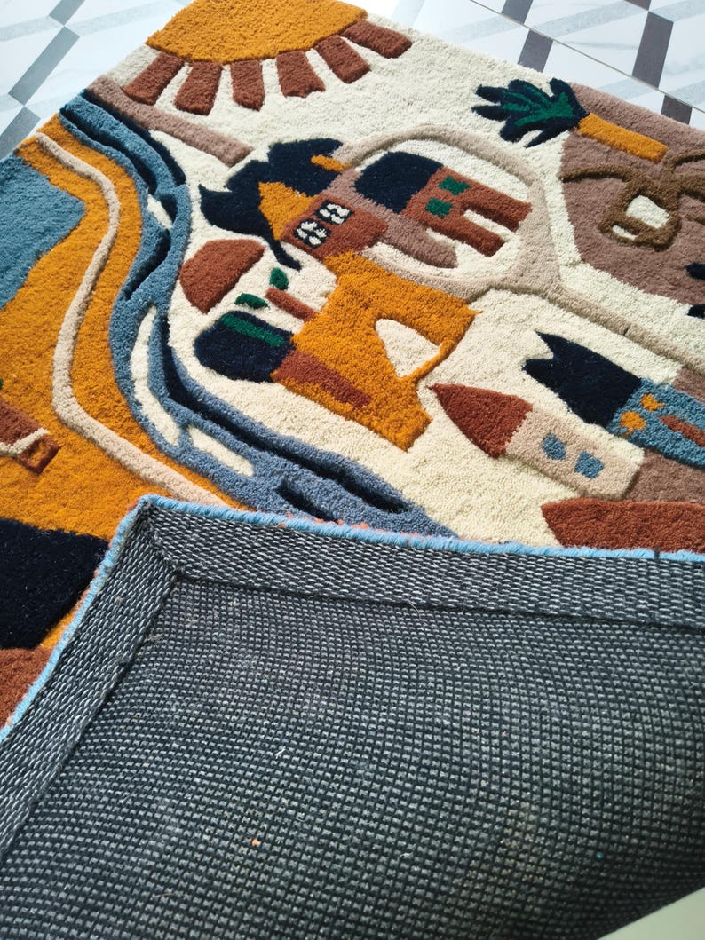 May include: A tufted rug featuring a cream background with a colorful landscape design including a yellow sun, blue river, and brown buildings. The rug has a textured pile surface and a dark gray, grid-patterned non-slip backing visible at the bottom.