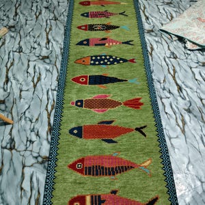 May include: A long, rectangular table runner in a vibrant lime green color featuring a repeating pattern of twelve stylized fish arranged in a vertical row. Each fish displays a unique geometric design and a distinct color palette of red, blue, and pink.