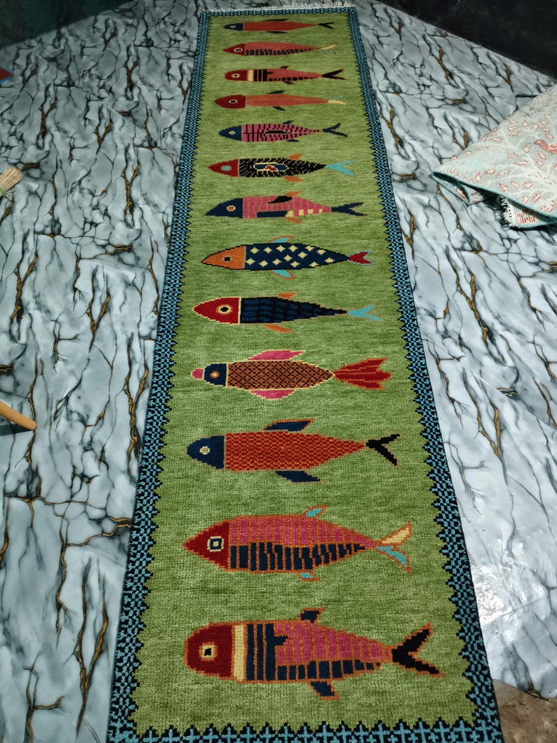 May include: A long, rectangular table runner in a vibrant lime green color featuring a repeating pattern of twelve stylized fish arranged in a vertical row. Each fish displays a unique geometric design and a distinct color palette of red, blue, and pink.
