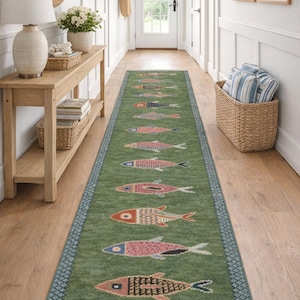 May include: A long, rectangular green runner rug featuring a repeating pattern of stylized fish in various colors including orange, pink, and blue. The rug has a dark blue geometric border and short white fringe along both of the narrow ends.