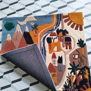 May include: A rectangular tufted rug featuring a colorful landscape illustration with mountains, a winding river, houses, trees, and a sun. The rug has a dark gray backing visible where one corner is folded over, revealing the textured surface.