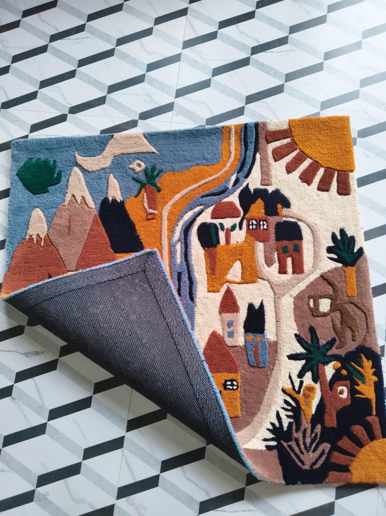 May include: A rectangular tufted rug featuring a colorful landscape illustration with mountains, a winding river, houses, trees, and a sun. The rug has a dark gray backing visible where one corner is folded over, revealing the textured surface.