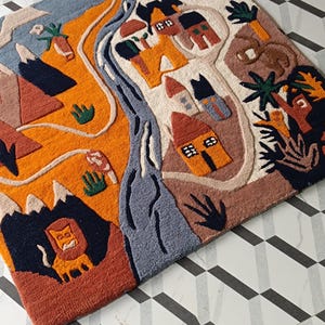 May include: A rectangular tufted rug featuring a vibrant landscape illustration with a bright orange sun, a winding blue river, stylized houses, mountains, and a lion. The design incorporates earthy tones of terracotta, cream, and dark blue.