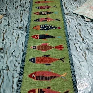 May include: A long, rectangular table runner in a light green hue featuring a vertical row of twelve stylized fish motifs. Each fish displays a unique geometric pattern and color combination, including shades of red, blue, orange, and pink. The runner is bordered by a dark blue and black patterned trim.