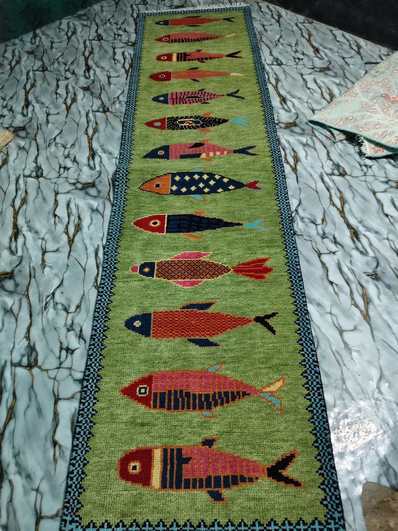 May include: A long, rectangular table runner in a light green hue featuring a vertical row of twelve stylized fish motifs. Each fish displays a unique geometric pattern and color combination, including shades of red, blue, orange, and pink. The runner is bordered by a dark blue and black patterned trim.