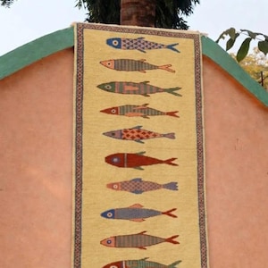 May include: A rectangular woven wall hanging featuring a light yellow background with a vertical row of ten stylized fish. Each fish displays a unique pattern and color combination, including shades of blue, red, green, and orange. The piece has a decorative border.